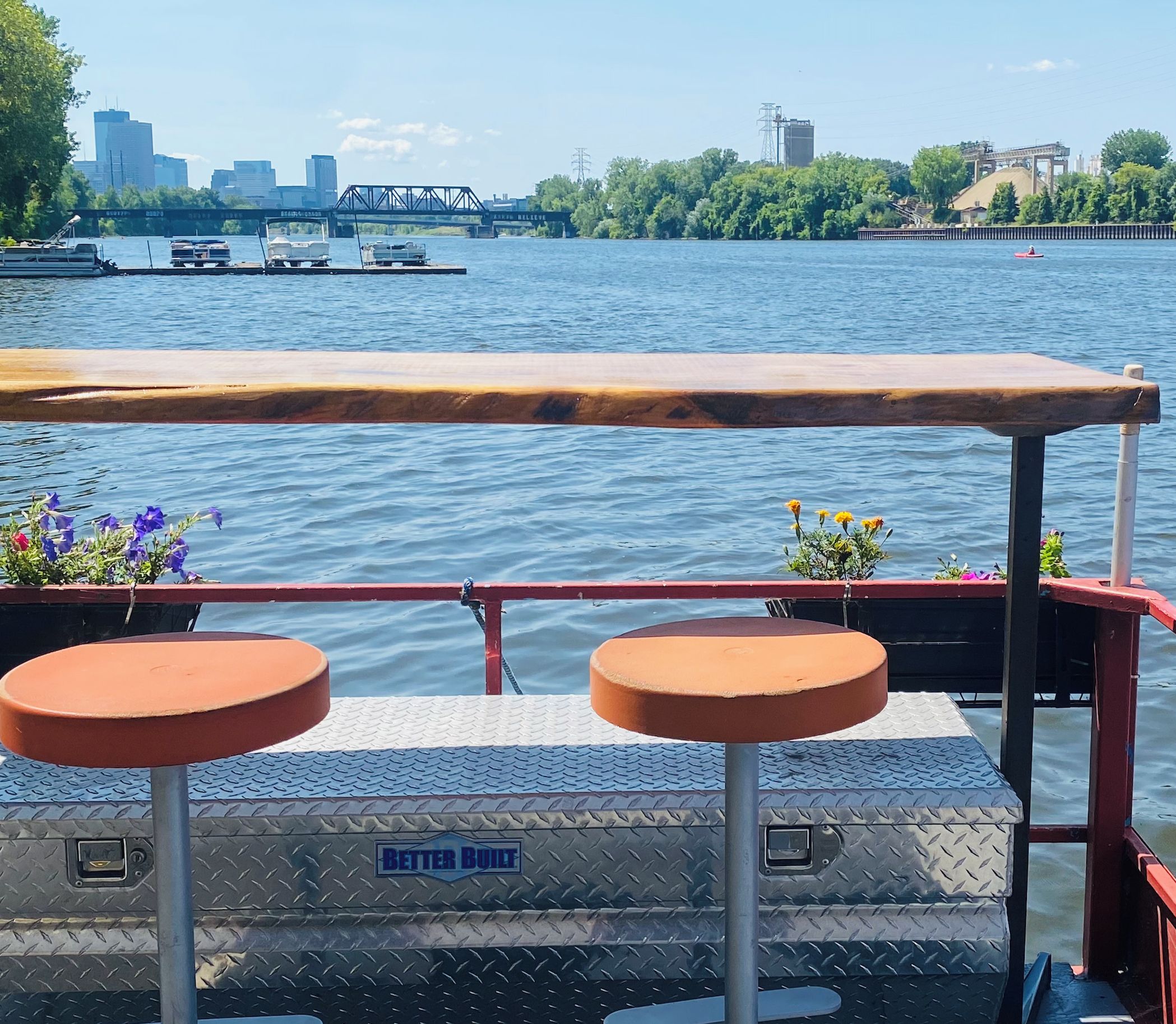 A boat with a metal table and two stools, a metal box labeled 'Better Built,' and a view of a city skyline across the water.