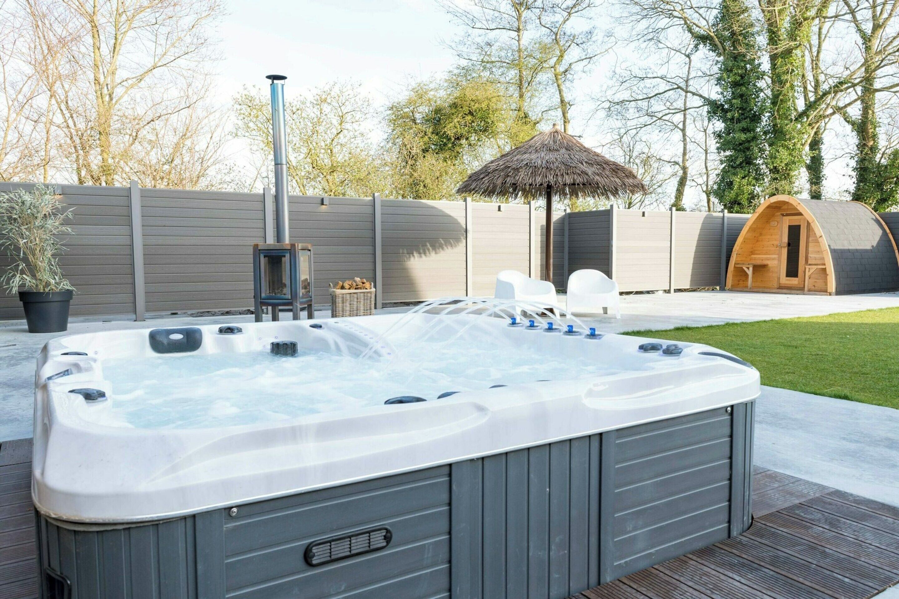 A hot tub with a wooden deck, a thatched umbrella, and a wooden cabin in the background.