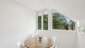 Quarto com mesa de madeira redonda, cadeiras brancas e amplas janelas com vista para árvores.
