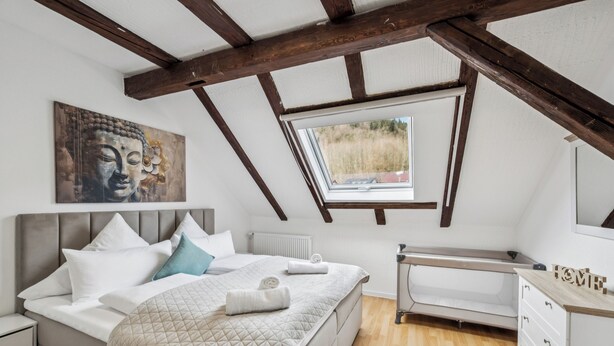 Quarto com teto inclinado, cama com almofadas, mesa de cabeceira, janela e um quadro na parede.