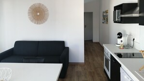Cozinha moderna com máquina de lavar louça, micro-ondas e bancada branca. Um sofá preto está em primeiro plano, e uma mesa de jantar branca é visível.