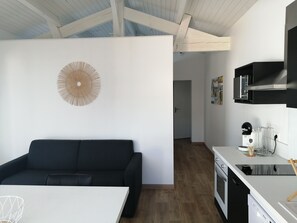 Eine moderne Küche mit Geschirrspüler, Mikrowelle und einer weißen Arbeitsplatte. Ein schwarzes Sofa steht im Vordergrund, und ein weißer Esstisch ist zu sehen.