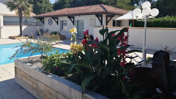 Uma área à beira da piscina com um deck de azulejos, um vaso de madeira com flores e um edifício com telhado e janelas em azulejo.