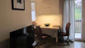 Ein modernes Wohnzimmer mit einem Flachbildfernseher, einem Esstisch mit Stühlen, einer Salatschüssel und einem großen Fenster mit Vorhängen.