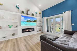 A living room with a TV, fireplace, and a view of a tropical beach. - 31214 Mimi lane (Frankford)