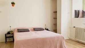 A bedroom with a bed, bedside tables, a hanging plant, a radiator, and a shelf unit. - Modern Apartment 2 in Historic Abbey (Cadier en Keer)