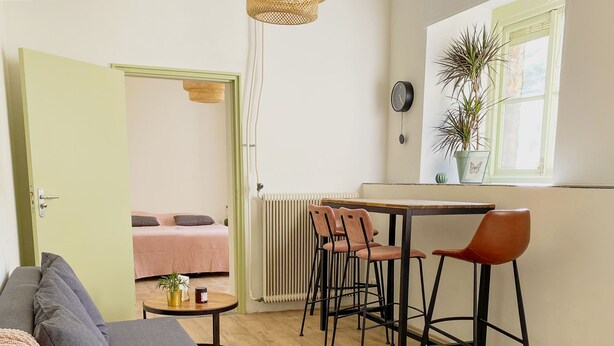 A compact living space with a kitchenette, a dining area with chairs, a sofa, a small table with a plant, and a radiator. - Modern Apartment 2 in Historic Abbey (Cadier en Keer)