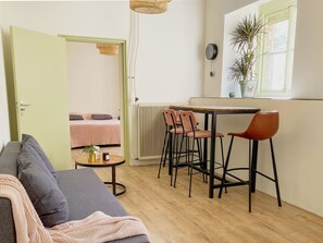 Un espacio habitable compacto con kitchenette, un área de comedor con sillas, un sofá, una pequeña mesa con una planta y un radiador.