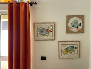 Une pièce avec un rideau, trois tableaux représentant des poissons encadrés accrochés au mur et un interrupteur.