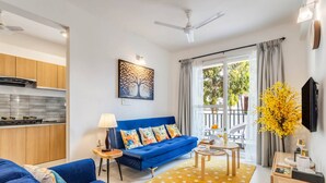 A modern living room with a blue sofa, a dining table, a kitchen area, and a balcony with a view. - Prime Candolim 2bhk w Pool Near Beach Nightlife (Candolim)