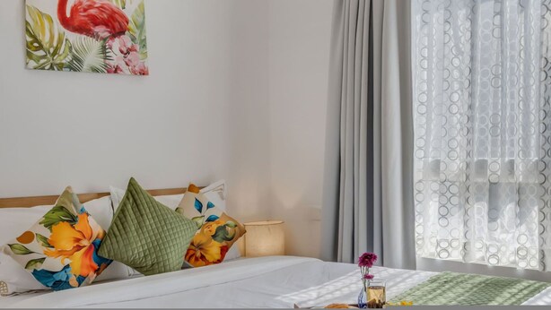 A hotel room with a bed, pillows, a nightstand, a lamp, and a painting of a flamingo. - Prime Candolim 2bhk w Pool Near Beach Nightlife (Candolim)