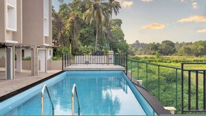 A swimming pool with a metal railing, surrounded by greenery and a fence, with a building on one side. - Prime Candolim 2bhk w Pool Near Beach Nightlife (Candolim)