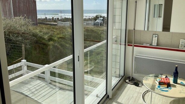 Een kamer met een grote schuifglazen deur naar een balkon met uitzicht op de oceaan en een houten gebouw.