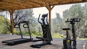 Ein Outdoor-Fitnessbereich mit Laufbändern, Crosstrainern und einem stationären Fahrrad unter einer Holzpergola, vor einer natürlichen Kulisse.