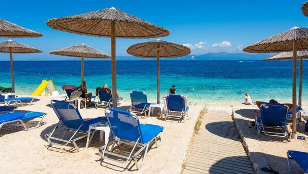 Ein Strand mit Liegestühlen und Reetsonnenschirmen, einem klaren blauen Meer und einem Holzsteg.