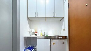 Cocina compacta con armarios blancos, cafetera, refrigerador pequeño y un cesto de basura azul.