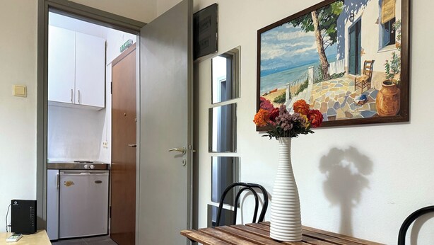 Un área de comedor con una mesa y sillas de madera, un jarrón blanco con flores, y un cuadro de una escena costera en la pared.