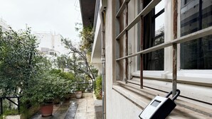 Un estrecho pasillo exterior con una ventana a la derecha, barandilla metálica y plantas en macetas.