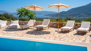 A poolside area with lounge chairs and umbrellas, a stone-tiled floor, and a view of the sea and mountains. - Sunrise Villa With 3 Bedrooms and 3 Bathrooms (Kefalonia)