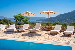 Ein Poolbereich mit Liegestühlen und Sonnenschirmen, ein Boden aus Steinplatten und Blick auf Meer und Berge.