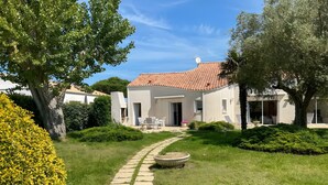 Une villa avec un toit en tuiles, un chemin pavé et un jardin agrémenté d’arbustes et d’un arbre.