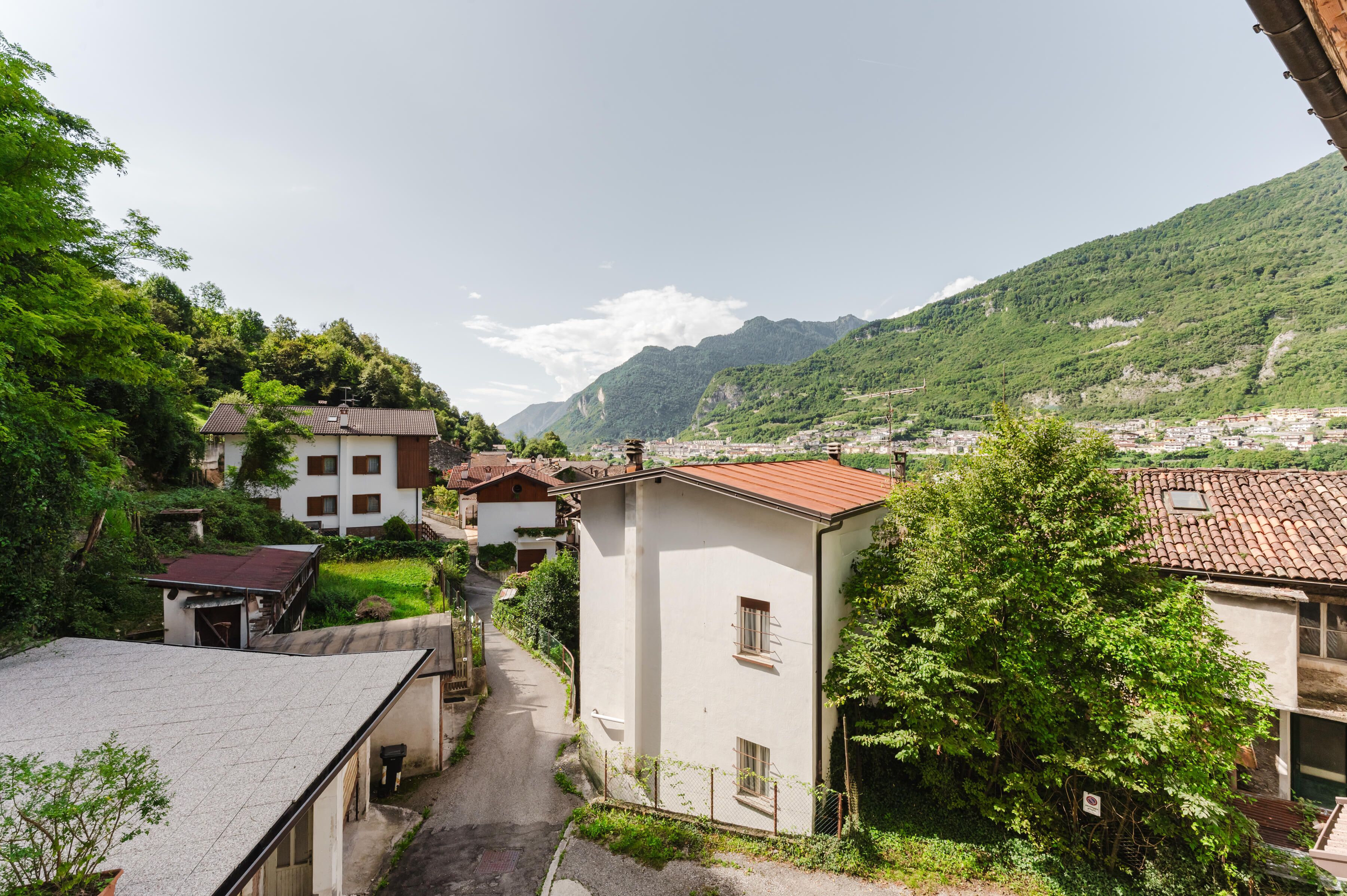 A narrow street in a mountainous village with traditional buildings, a clear sky, and lush greenery.