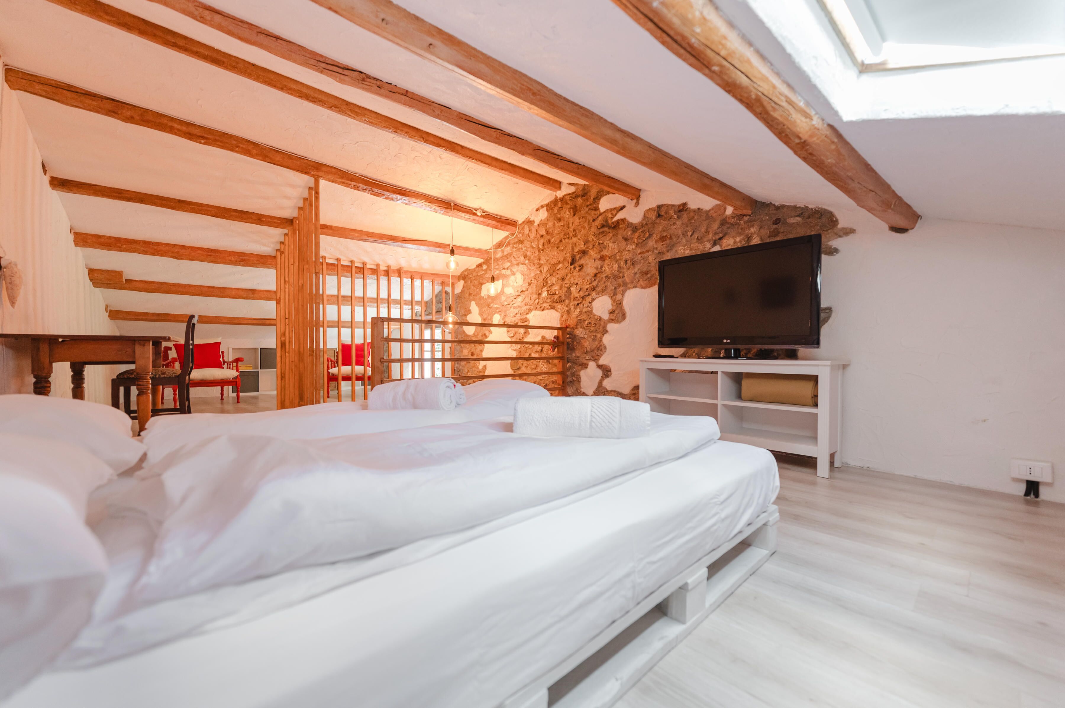 Une chambre à coucher avec un lit, des poutres en bois, un mur en pierre, une télévision et un bureau avec des chaises.