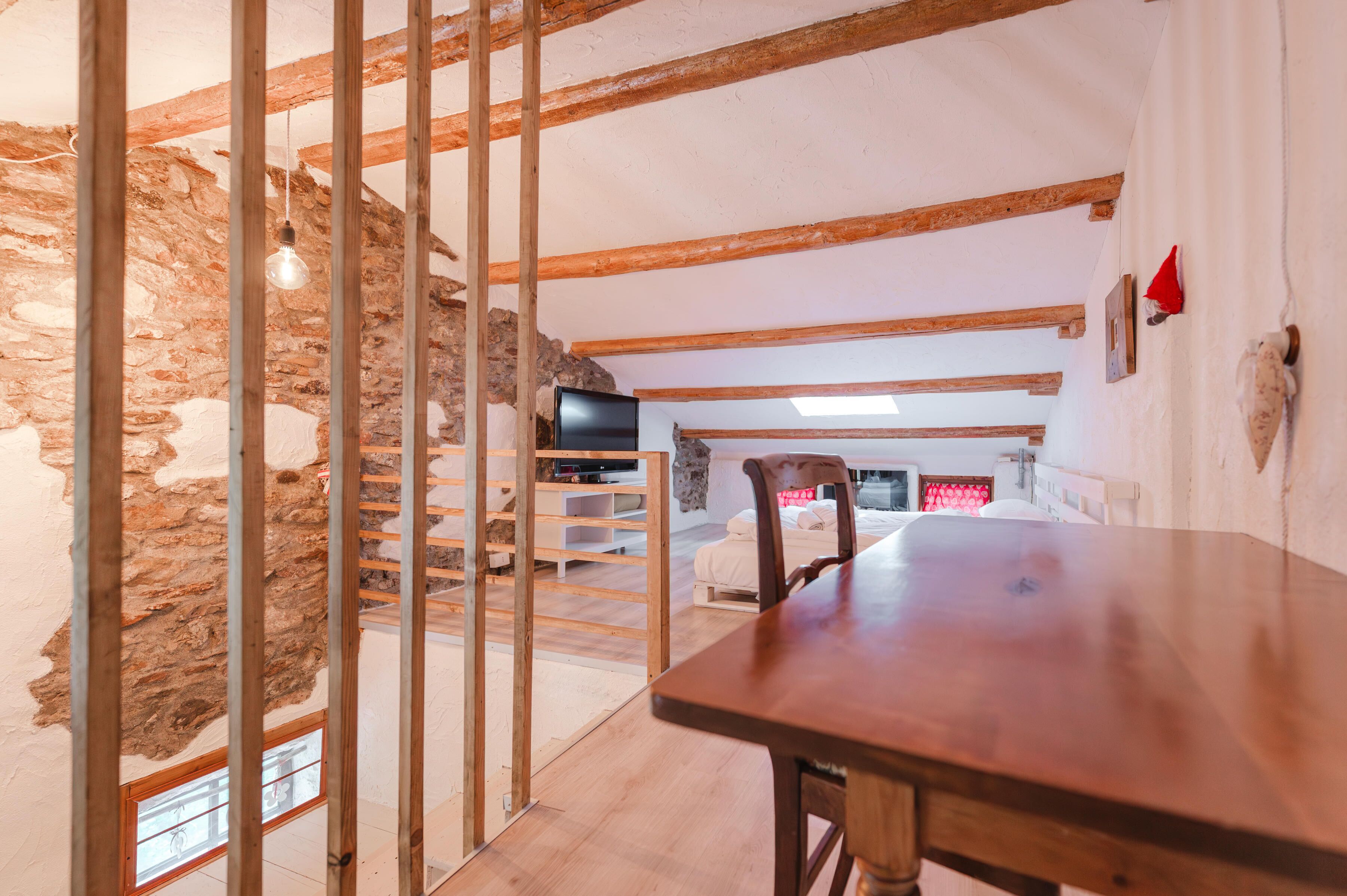 A room with exposed wooden beams, a stone wall, a wooden table, and a television in the background.