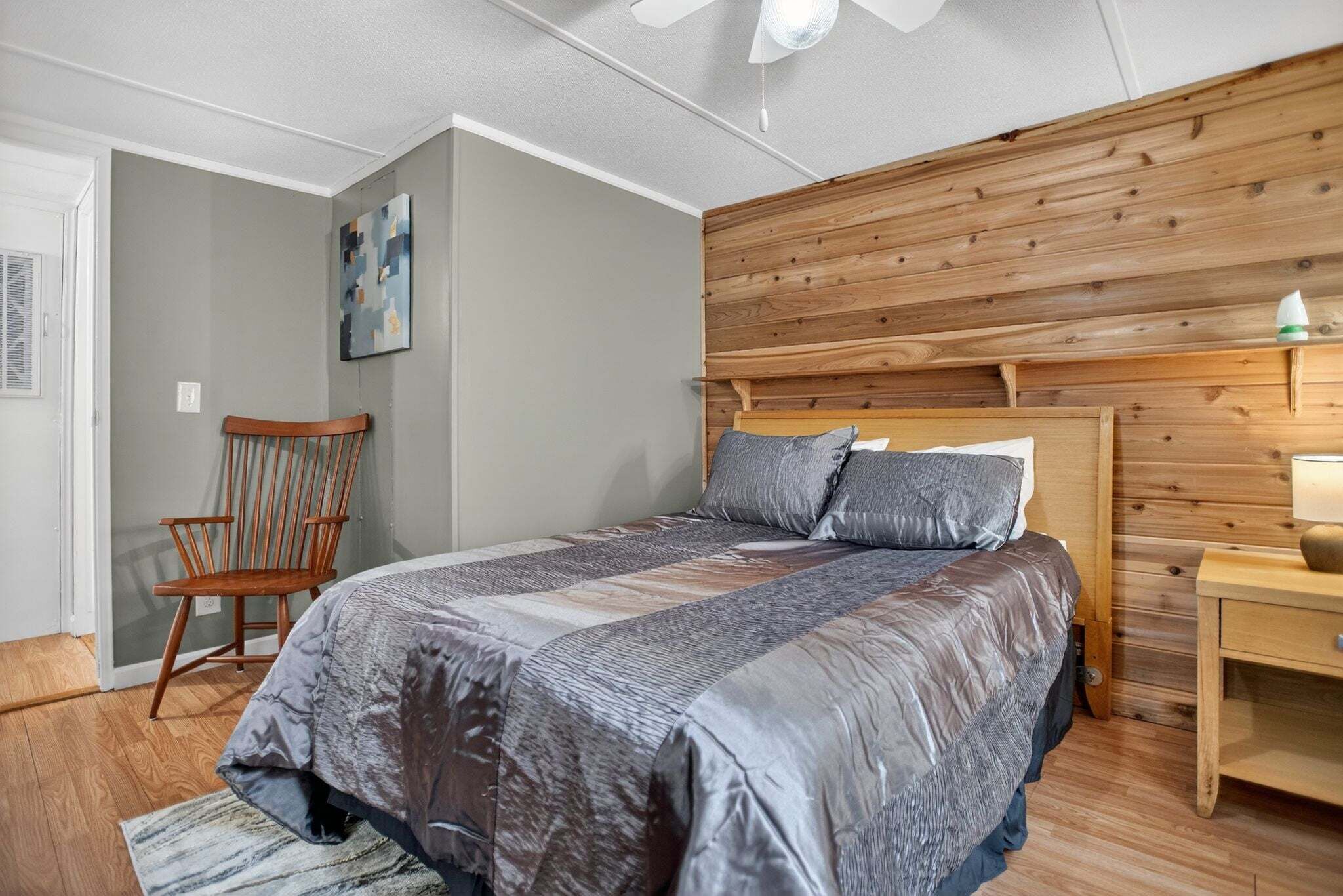 Une chambre à coucher avec une tête de lit en bois, un lit avec une literie grise, une chaise en bois et une table de chevet avec une lampe.