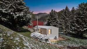 A small, single-story house with a red barn in the background, surrounded by snow-covered trees and a hilly landscape. - Peaceful Pines - 2br Getaway Near Blue Ridge 2 Bedroom Home (Burnsville)