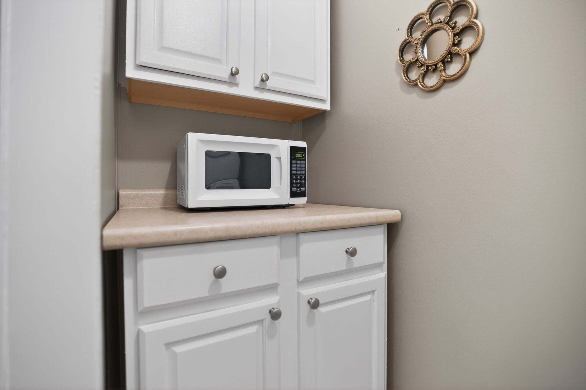 A white microwave on a beige countertop, white cabinets, and a decorative metal piece on the wall.