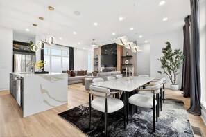 Una cocina moderna con una isla central, una zona de comedor con mesa y sillas blancas, y un espacio de sala de estar con sofá y televisor.