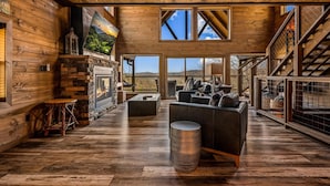 A spacious living area with a fireplace, wooden flooring, and a large window overlooking a scenic landscape. - Kelly's Escape Cabin (Sevierville)