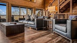 A spacious living room with a wooden ceiling, large windows, a sofa, a coffee table, and a staircase. - Kelly's Escape Cabin (Sevierville)