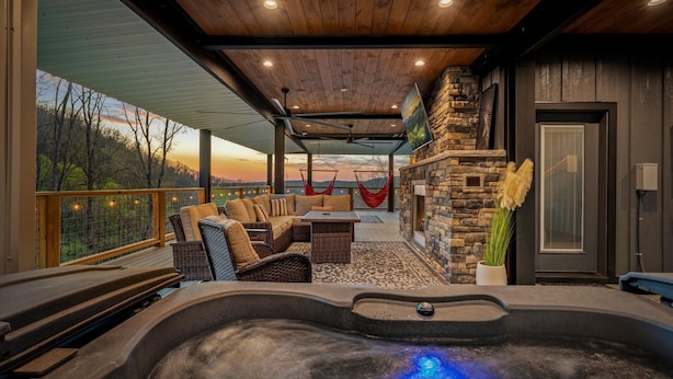 A hot tub with a blue light, wicker furniture, a stone fireplace, and a view of trees. - Kelly's Escape Cabin (Sevierville)