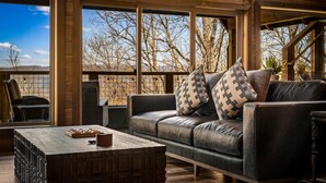A cozy living area with a leather sofa, wooden coffee table, and large windows offering a view of trees and a clear sky. - Kelly's Escape Cabin (Sevierville)