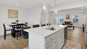 A modern kitchen with a central island, dining area with chairs, and a living room with a sofa and windows. - New 4br Townhome Near Harpers Ferry 4 Bedroom Townhouse (Charles Town)