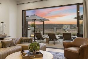 A modern living room with a leather sofa, armchairs, and a coffee table. There is a large sliding glass door leading to an outdoor patio with a view of the hills. - Ever-changing Sky 7 Bedroom Home (Washington)