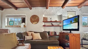 A cozy living room with a sofa, a coffee table with a magazine, a television displaying a beach scene, and a bookshelf. - Casita Adobe Duplex (Grand Junction)
