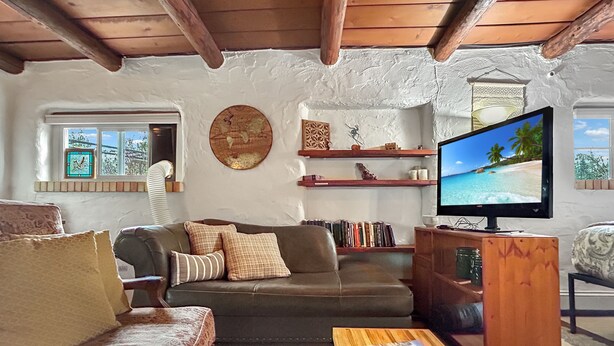 A cozy living room with a sofa, a coffee table with a magazine, a television displaying a beach scene, and a bookshelf. - Casita Adobe Duplex (Grand Junction)