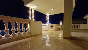 Een terras buiten, 's nachts verlicht, met betegelde vloer, witte balustrades en meerdere plafondlampen.
