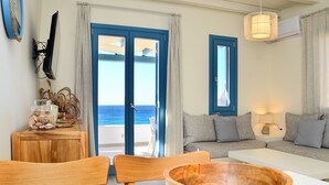 A room with a wooden dining table, a sofa, a TV mounted on the wall, and a view of the sea through the doors.