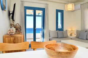 A room with a wooden dining table, a sofa, a TV mounted on the wall, and a view of the sea through the doors.