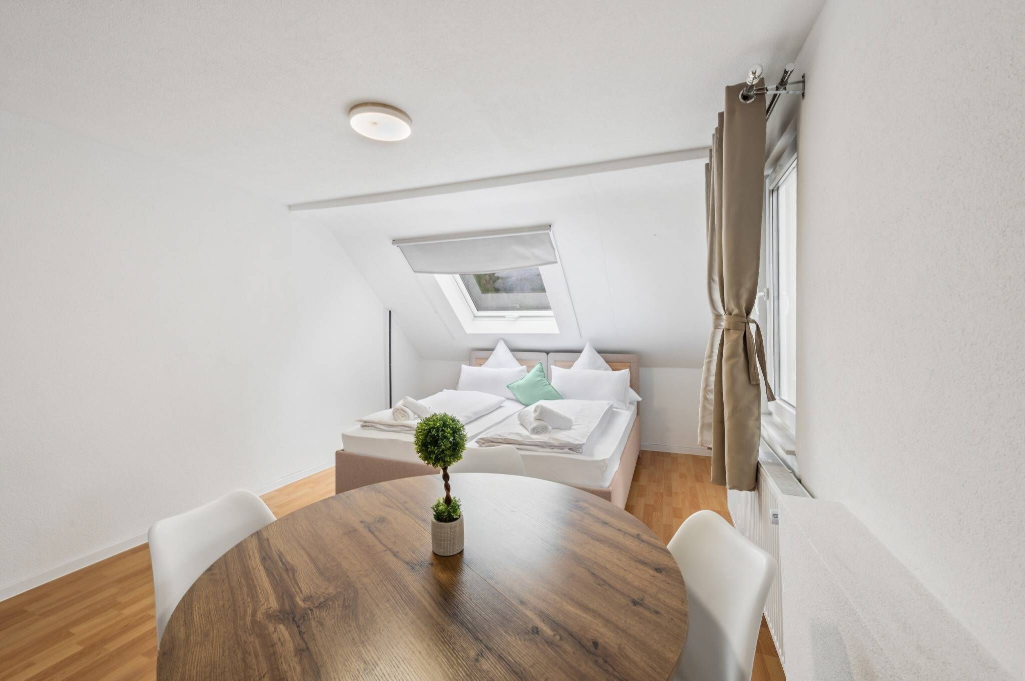 Une chambre moderne avec une lucarne, un lit recouvert de draps blancs, une table ronde en bois ornée d’une plante en pot et des chaises blanches.