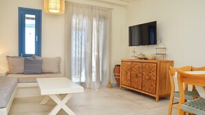 A living room with a sofa, a white coffee table, a wooden cabinet, a TV mounted on the wall, and a window with curtains. - Villa Amorgos, Sohoro Koufonisia (Koufonisia)