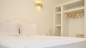 A hotel room with a bed, a wall-mounted light, a white fan, and a built-in wardrobe with hangers. - Villa Amorgos, Sohoro Koufonisia (Koufonisia)