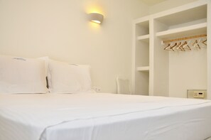 Une chambre d’hôtel avec un lit, une lampe fixée au mur, un ventilateur blanc et une armoire intégrée avec des cintres.