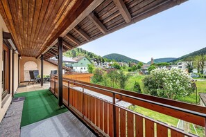 En balkon med gelænder af træ, en grøn måtte og udsigt til huse og bakker.