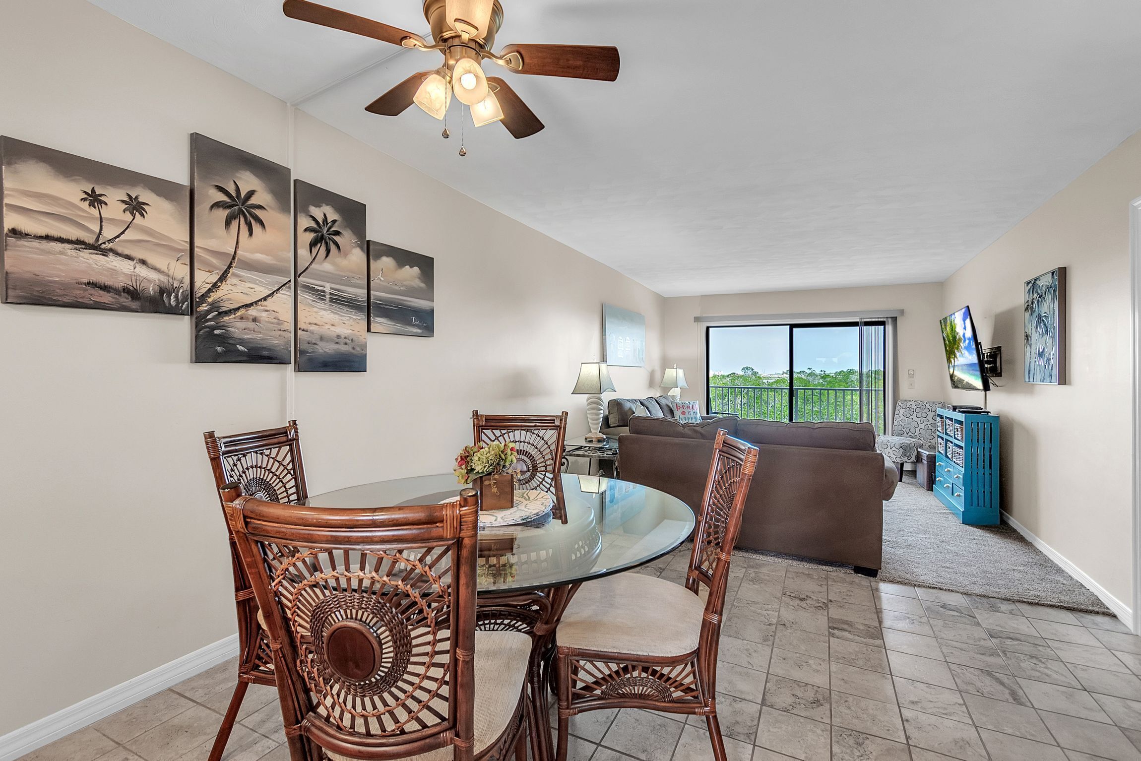 A dining area with a glass table and wicker chairs, a ceiling fan, and artwork on the walls.