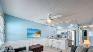 Sala de estar com televisão, ventilador de teto, cozinha e um banco de bar.